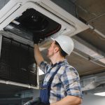 hvac technician wearing hardhat and holding screwdriver, repairing cassette type air conditioner unit on ceiling