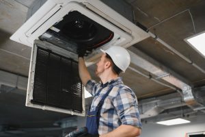 hvac technician wearing hardhat and holding screwdriver, repairing cassette type air conditioner unit on ceiling
